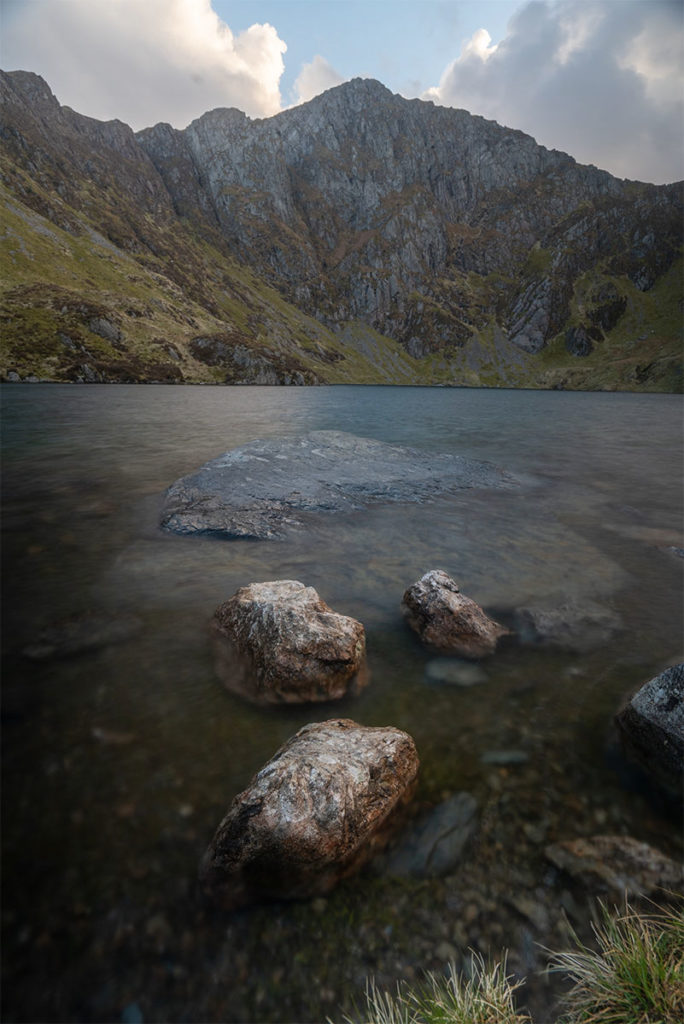 The glacial lake on Cader Idris mountain with three grey rocks in the foreground and mountains in the background. There are some grey and white clouds in the sky above the mountain. 