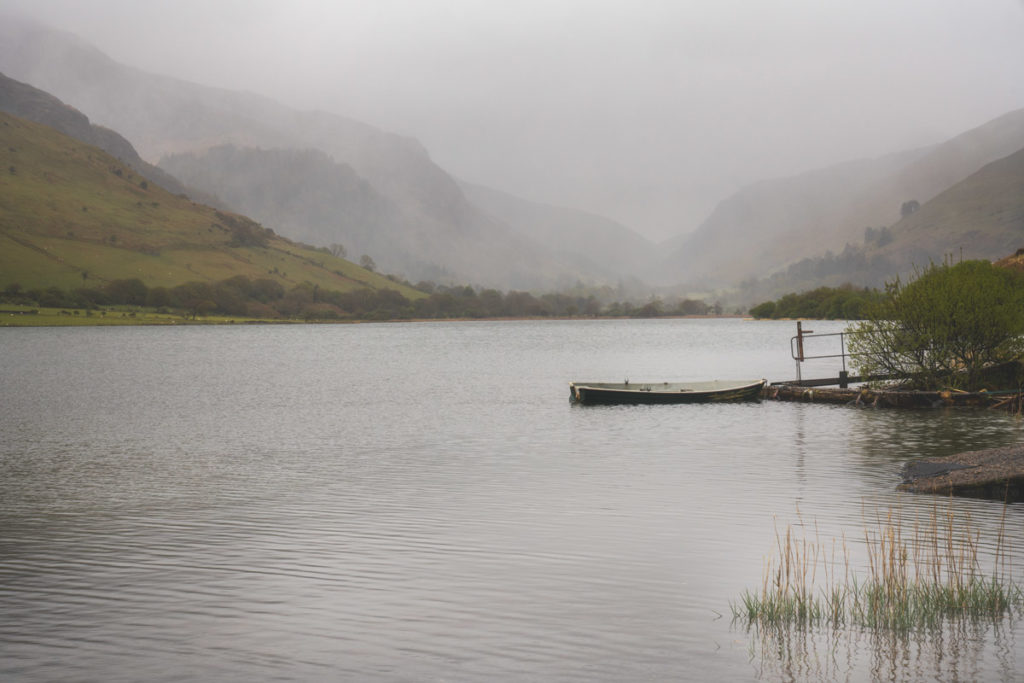  Still Lake with foggy background and green mountains on either side of the frame. There is a small wooden boat to the right tied up next to a ramp leading to it. 