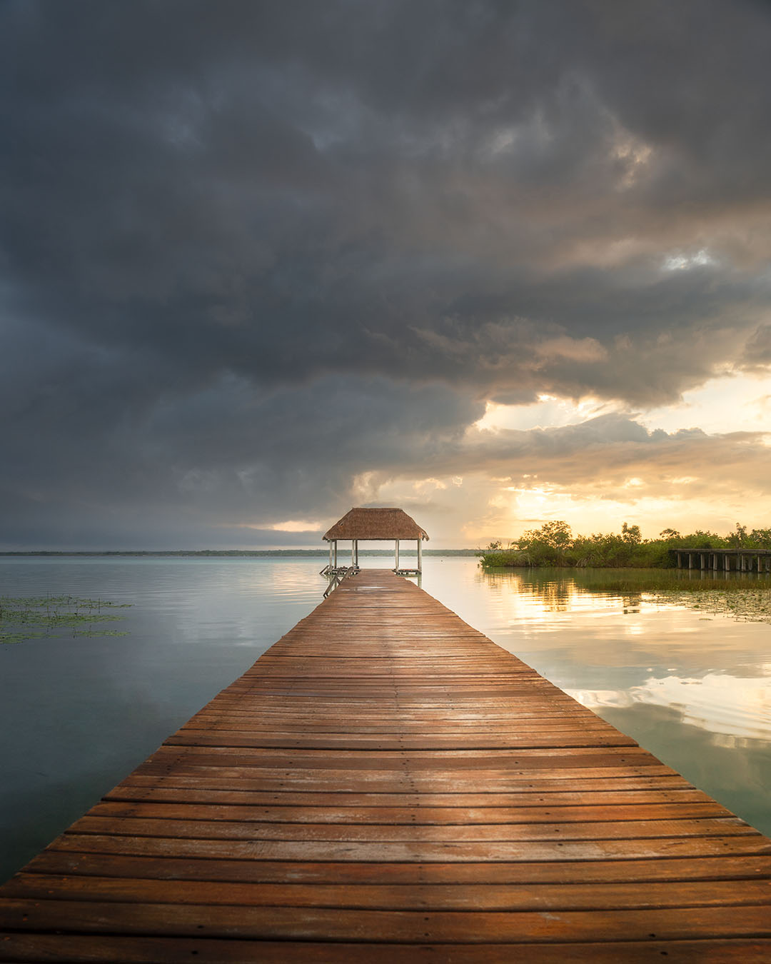 A photograph taken on a cloudy day of a long boardwalk into the ocean with a thatched roof open palapa at the end with dark grey clouds in the sky