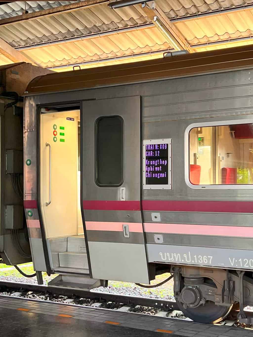 The grey sleeper train from Bangkok to Chiang Mai with pink bands across it and the door is open with a small digital sign saying "Train 009 Car 12 Krung Thep Aphi Wat Chiang Mai"