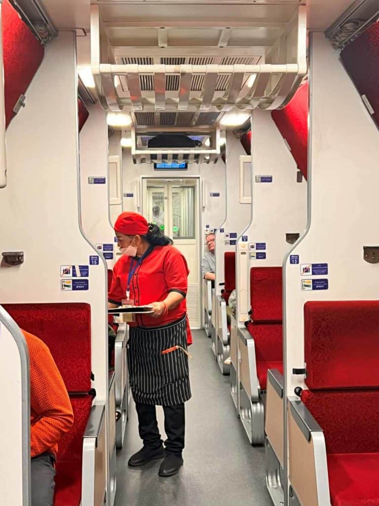 The 2nd class AC cabin on the sleeper train Bangkok to Chiang Mai which is a row of red seats and a lade wearing all red with black trousers taking orders for food