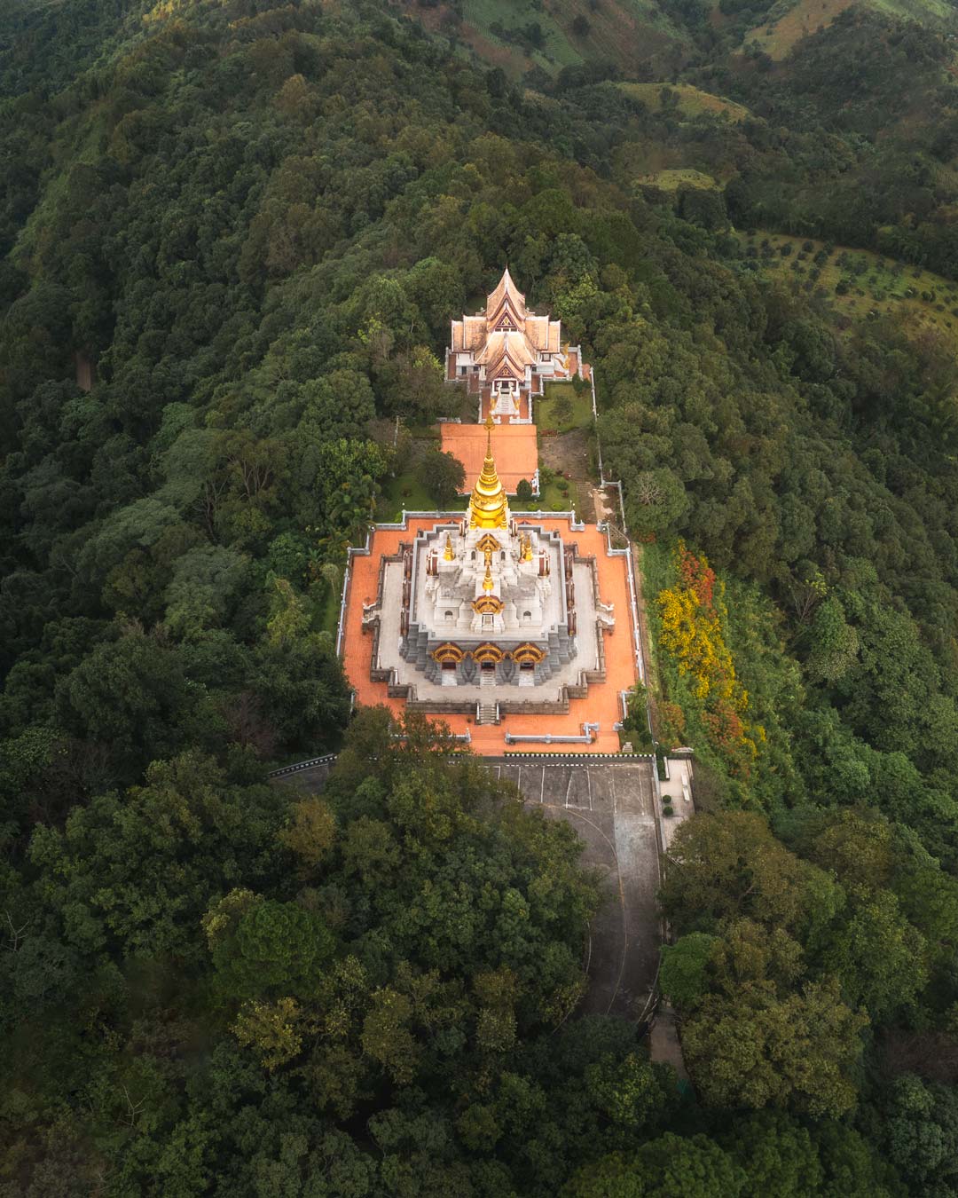 A drone shot of Wat Chedi Srinagarindra Stit Maha Santi Khiri Temple - a peach, white and gold coloured temple taken from above surrounded by green trees and hills
