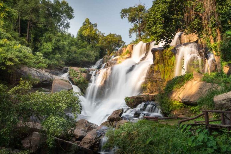 A professional landscape photo of Mae Klang Waterfall, featuring a silky, long-exposure effect on the cascading water, framed by vibrant green foliage and rocky outcrops with golden light shining on to the falls