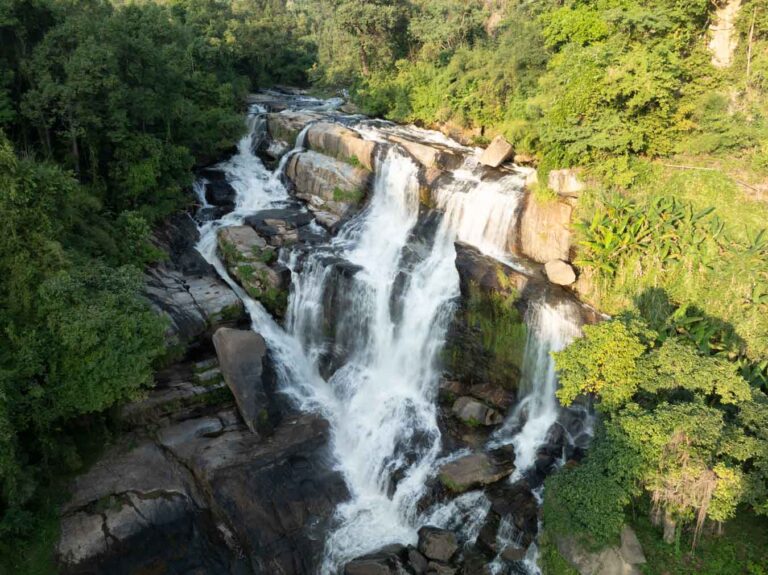 A dramatic drone photo showcasing Mae Klang Waterfall in Doi Inthanon National Park, with its tiered cascades flowing powerfully through lush greenery under soft, natural light. The waterfall is very wide across lots of rocks.