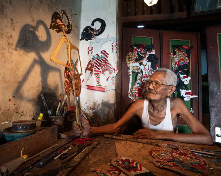 An elderly artisan sits at a wooden workspace surrounded by colorful tools and handcrafted shadow puppets. He holds up one intricately designed puppet, its shadow cast dramatically on the wall behind him. The scene is illuminated by warm light, highlighting his focus and the rich cultural details of his craft.