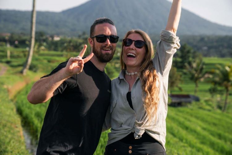 Emma and Steffan from Inwanderment doing the peace sign and one hand in the air with a rice terrace and mountain the background smiling at the camera