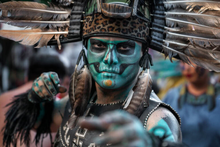 A Mayan dancer in Valladolid stares intensely into the camera, his face painted in striking turquoise and black designs resembling a skull. He wears an elaborate headdress adorned with large feathers and a leopard-print band, with additional feathers and beads hanging from his costume. His outstretched hand, also painted, reaches forward in a dynamic pose. The blurred background of other performers and festival-goers captures the energy of the cultural celebration.