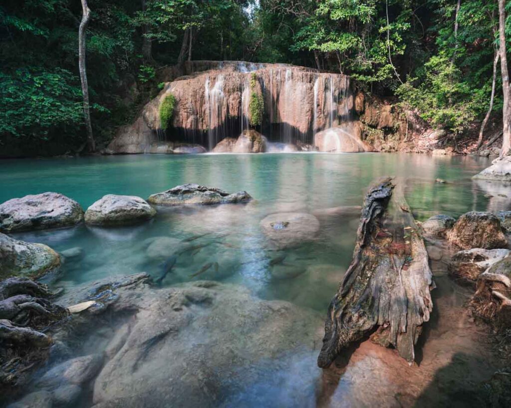A wide-angle view of one of the lower tiers of Erawan Waterfalls in Kanchanaburi, Thailand, featuring a turquoise pool with smooth rocks and a weathered log in the foreground, surrounded by lush green jungle and a curtain of gentle cascades flowing over limestone.