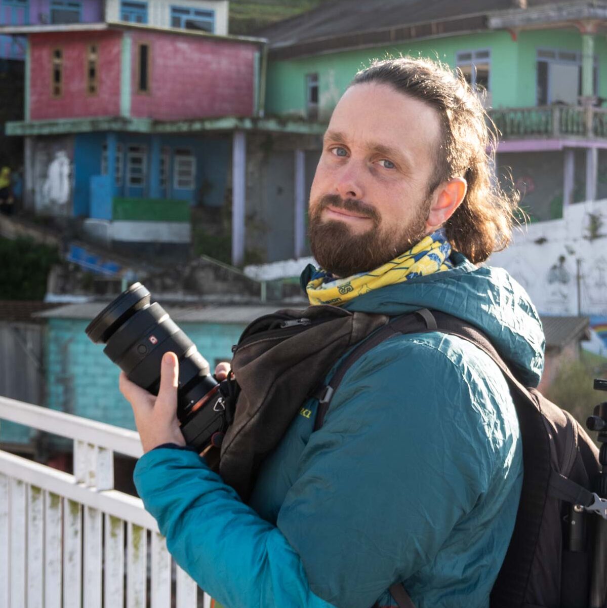 Steffan, a bearded man with long, curly hair tied back, stands on a white wooden bridge holding a professional camera with a large lens. He wears a blue and green insulated jacket, a yellow bandana around his neck, and carries a black backpack with photography gear. Behind him, colorful houses in shades of pink, green, and blue are built on a hillside, with bright natural light casting a warm glow on the scene.
