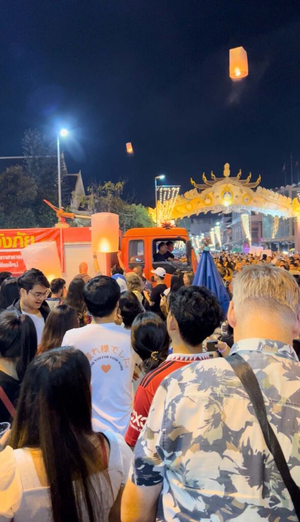 A large crowd gathers in the street during the lantern festival in Chiang Mai as floating lanterns rise into the sky. A fire engine is present in the scene, highlighting safety concerns amid the dense crowd.