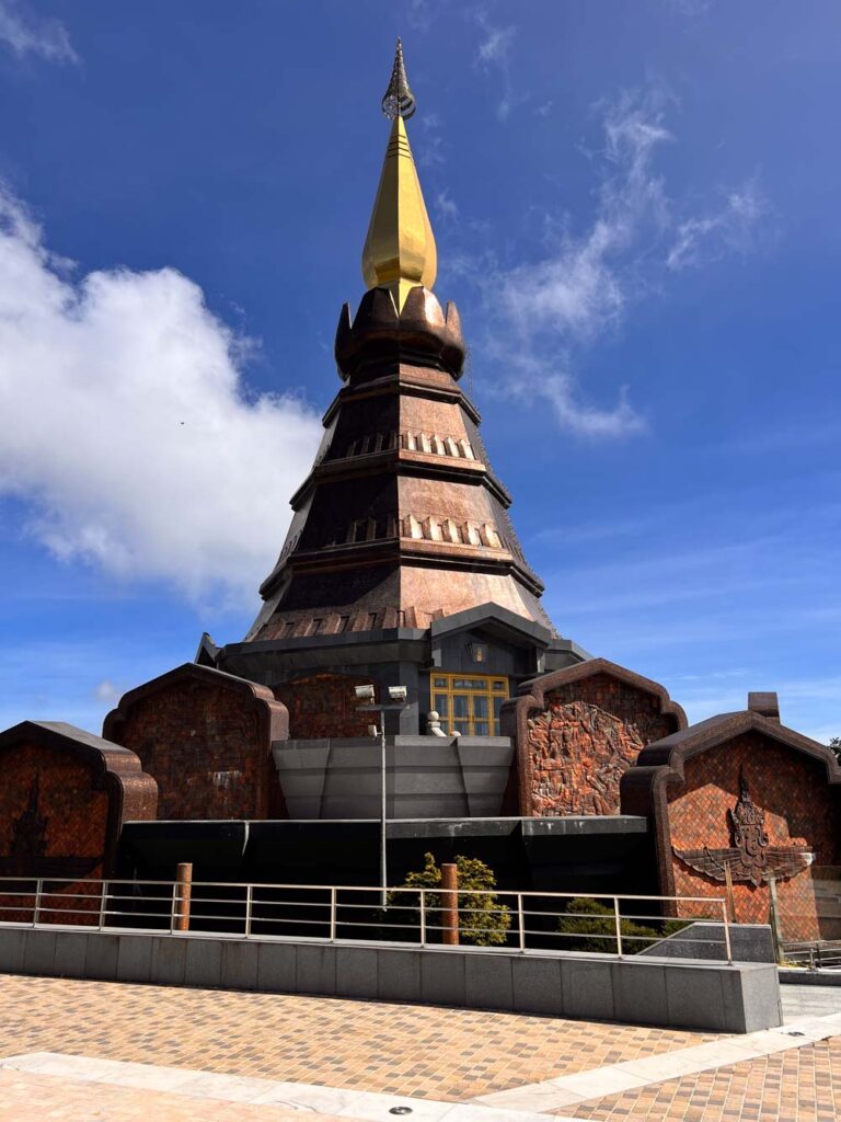 Close-up of the Kings Pagoda, one of the two Royal Pagodas at Doi Inthanon National Park in northern Thailand, featuring dark brown and bronze-toned stonework with a golden spire. The sky is bright blue with scattered clouds, and intricate carvings decorate the base, highlighting the unique architectural details.