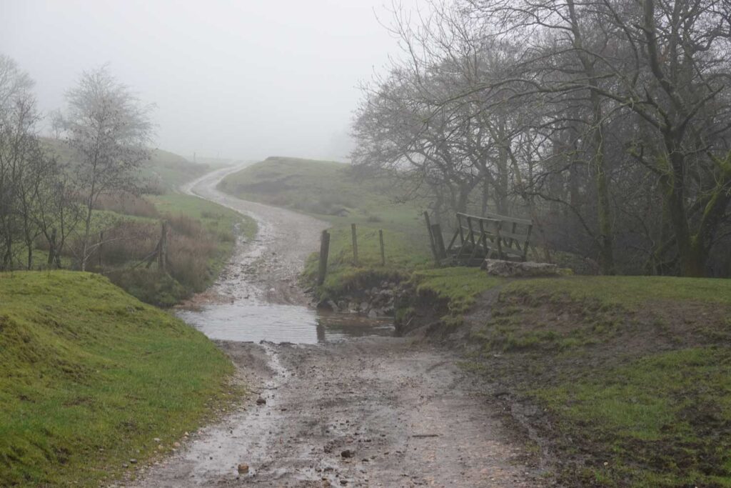 Misty countryside track near Hollinsclough, where the route to Chrome Hill walks crosses a shallow stream beside a wooden footbridge
