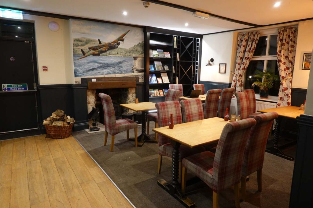Cozy dining area inside the Ladybower Inn with wooden tables and red plaid upholstered chairs arranged around a stone fireplace. A wall mural of a historic aircraft flying over a reservoir hangs above the fireplace, with bookshelves, patterned curtains, and warm lighting creating a welcoming countryside pub atmosphere near Derwent Edge.
