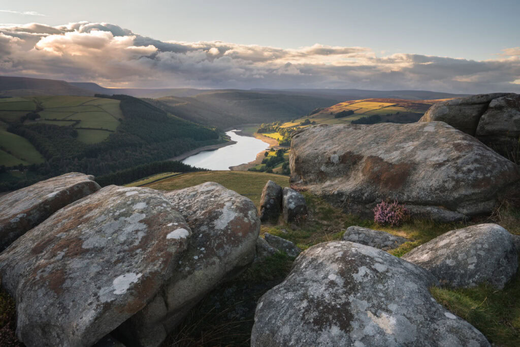 Large weathered gritstone boulders on Derwent Edge overlook a winding reservoir and patchwork fields in the Upper Derwent Valley. The late August light breaks through dramatic clouds, illuminating green hills and woodland below the viewpoint at Whinstone Lee Tor.
