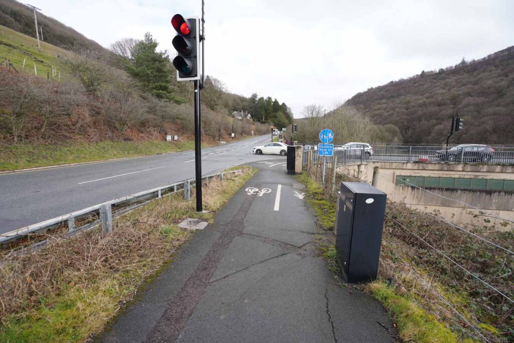 A shared cycle path runs alongside a busy road with a red traffic light overhead and white bicycle symbols painted on the tarmac. Cars wait at the junction ahead, and a blue sign indicates the designated route for cyclists and pedestrians through the valley.