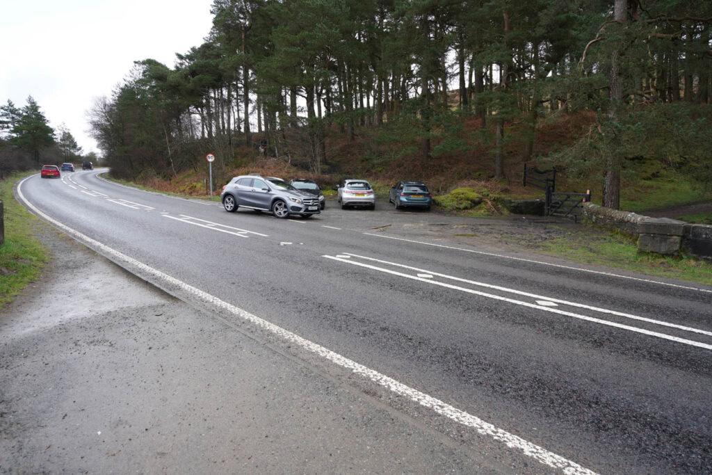 Parked cars beside a roadside pull off next to a dense pine forest on a damp overcast day. This small parking area is commonly used as a starting point for the Derwent Edge walk near Ladybower Reservoir in the Peak District.