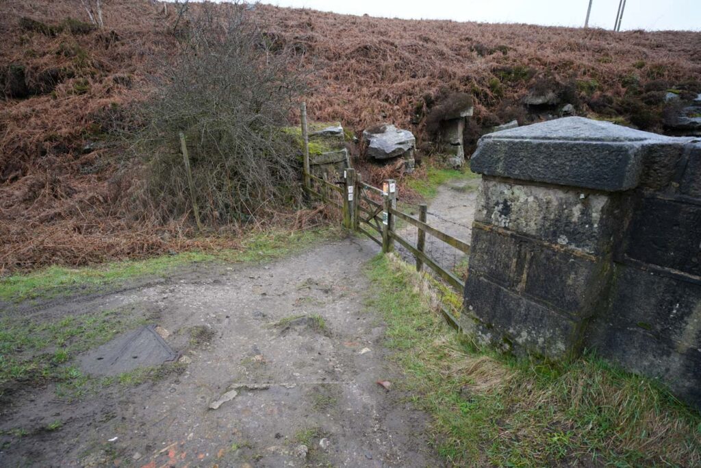 A wooden gate marks the start of a footpath leading onto open moorland with rocky outcrops and brown bracken. This path leads hikers onto Derwent Edge above Ladybower Reservoir in the Peak District.