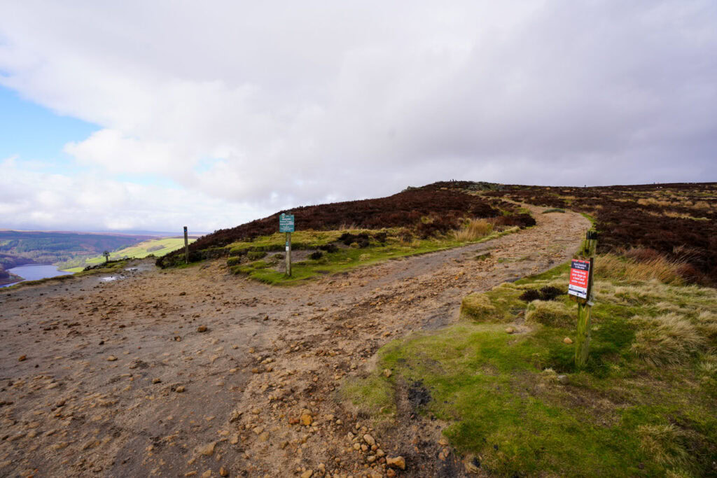 A rocky walking path splits across open moorland with heather and grass under a cloudy sky. Trail signs mark the route along Derwent Edge with views stretching across the hills and Ladybower Reservoir in the Peak District.