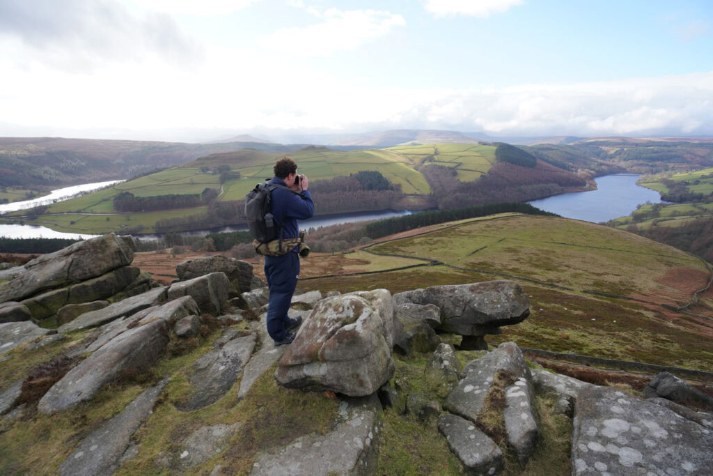 A hiker stands on rocky outcrops along Derwent Edge looking out across the wide valley below. Ladybower Reservoir winds through the landscape with green fields, forests, and rolling hills stretching across the Peak District.