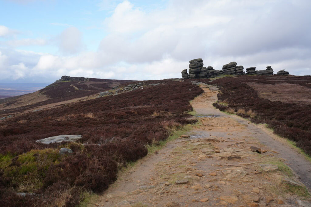 A rocky path winds through heather covered moorland toward stacked coach and horses rock formations along Derwent Edge. The distinctive stone towers rise above the landscape along this dramatic ridge in the Peak District.
