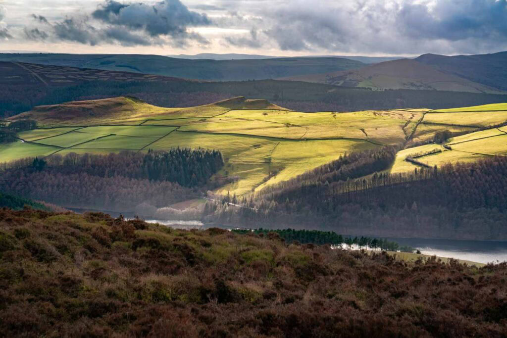 Winter landscape photography from Derwent Edge overlooking Crook Hill in the Peak District. Sunlight breaks through heavy clouds to illuminate bright green patchwork fields above the reservoir, with dark woodland and moorland in the foreground.