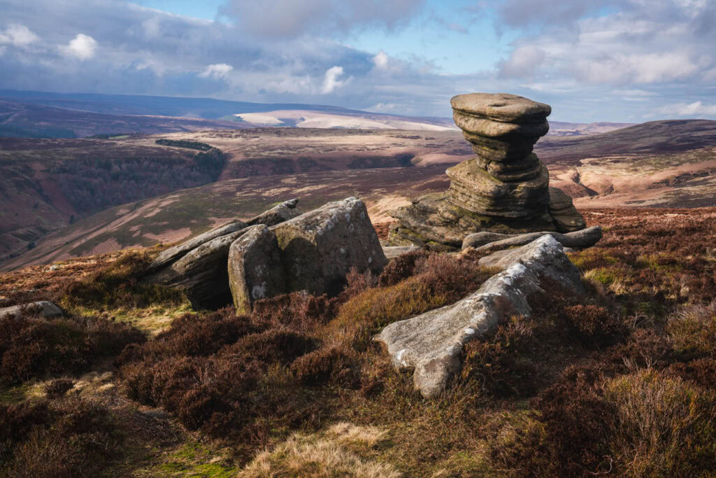 A distinctive stacked gritstone rock formation known as the Salt Cellar rises above the heather covered moorland on Derwent Edge. Rolling hills and wide open landscapes stretch across the Peak District under a dramatic cloudy sky.