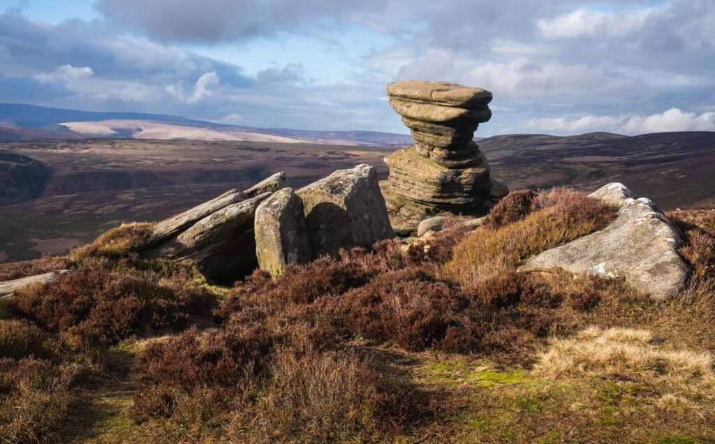 The Salt Cellar rock formation rises in stacked layers of gritstone above the heather covered moorland on Derwent Edge. The rugged Peak District landscape stretches into the distance beneath a partly cloudy sky.