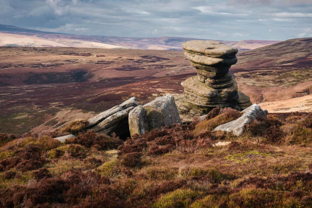 The Salt Cellar rock formation stands stacked above the heather covered moorland along Derwent Edge. Rolling hills stretch across the Peak District under dramatic clouds in the distance.