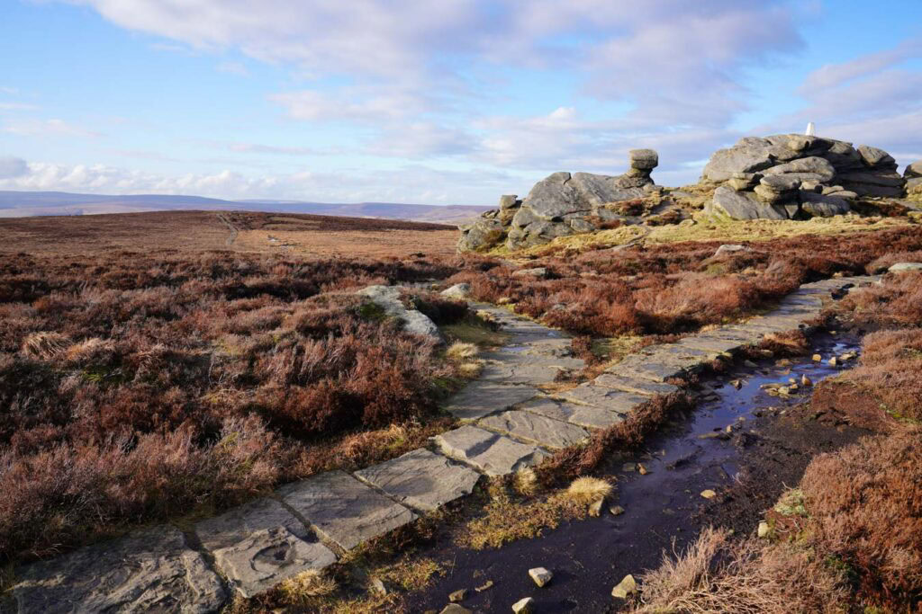 A stone slab path winds through wet heather moorland toward the rocky summit of Back Tor on Derwent Edge. Gritstone boulders rise above the ridge under a bright blue sky in the Peak District.