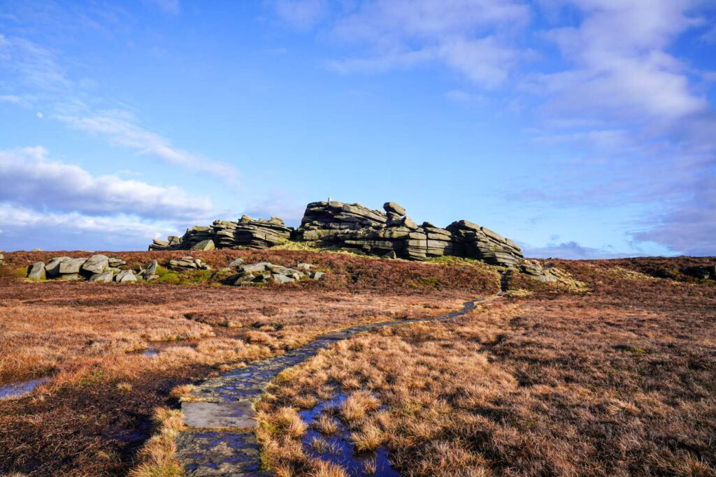 A stone slab path leads across the open moorland toward the rugged gritstone rocks of Back Tor on Derwent Edge. Brown grasses and heather stretch across the Peak District landscape beneath a bright blue sky.