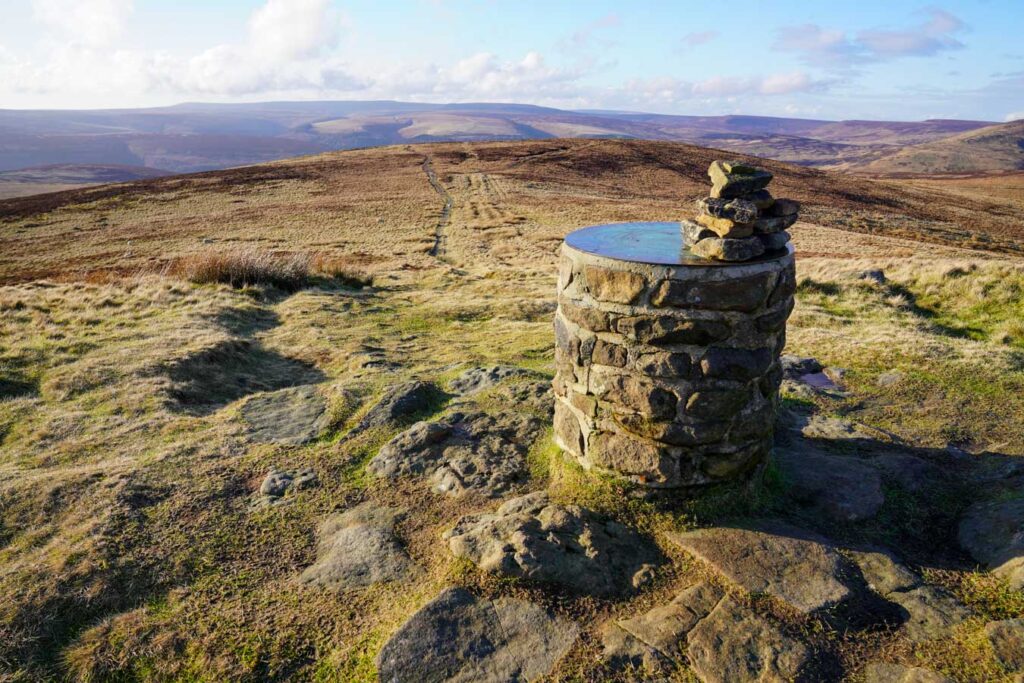 A stone summit marker with a small stack of rocks stands on the Lost Lad summit along Derwent Edge. The narrow trail continues across the wide open moorland with sweeping views over the Peak District.