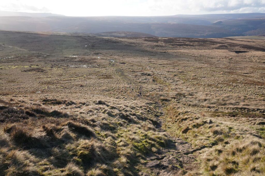 A narrow dirt path descends through rough grassy moorland along Derwent Edge. Rolling hills and wide open views stretch across the Peak District beneath soft sunlight.