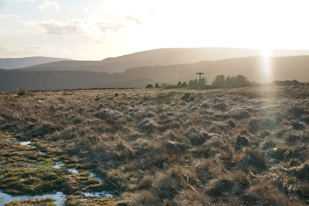 A wooden cross stands on open moorland along Derwent Edge with sunlight breaking through clouds over the distant hills. Frosted grasses and wide views stretch across the Peak District landscape.