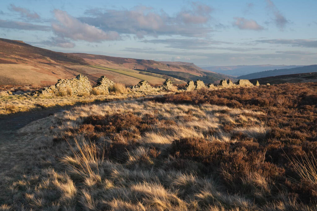 A landscape photography shot in the Peak District showing crumbling stone wall ruins stretching across heather covered moorland along Derwent Edge. Rolling hills and valleys extend into the distance beneath soft evening light.