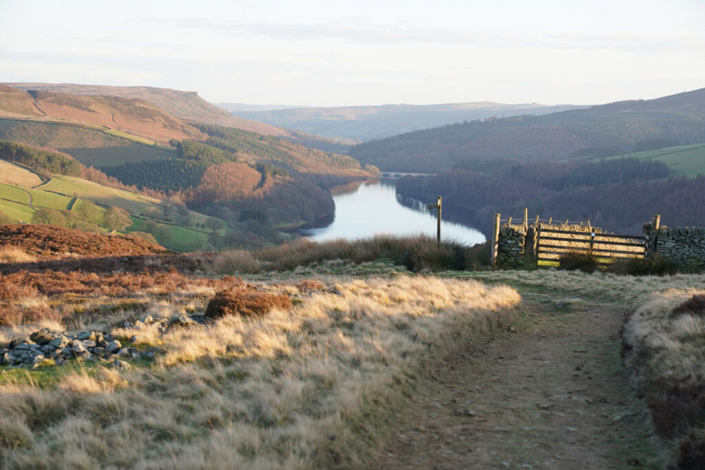 A landscape photography shot in the Peak District showing a dirt path leading toward a wooden gate above Ladybower Reservoir. Rolling hills, forests, and patchwork fields fill the valley below in soft golden light.