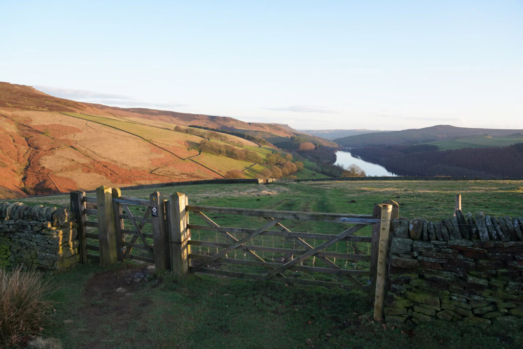 A wooden gate in a dry stone wall overlooks the winding waters of Ladybower Reservoir from Derwent Edge. Rolling hills, patchwork fields, and forests stretch across the Peak District valley in warm evening light.