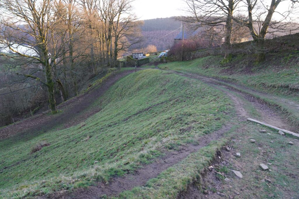 A dirt walking path splits across a grassy hillside beneath bare trees near the end of the Derwent Edge walk. A gate and small farm buildings sit ahead at the edge of the Peak District countryside.