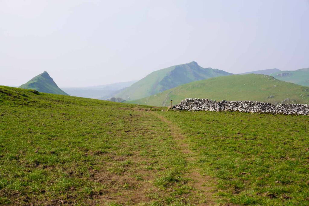 Rocky footpath descending toward Chrome Hill in the Peak District Derbyshire with green fields, a farm building, and dry stone walls below under soft hazy skies. The trail and surrounding hills showcase the varied terrain you can expect on chrome hill walks.