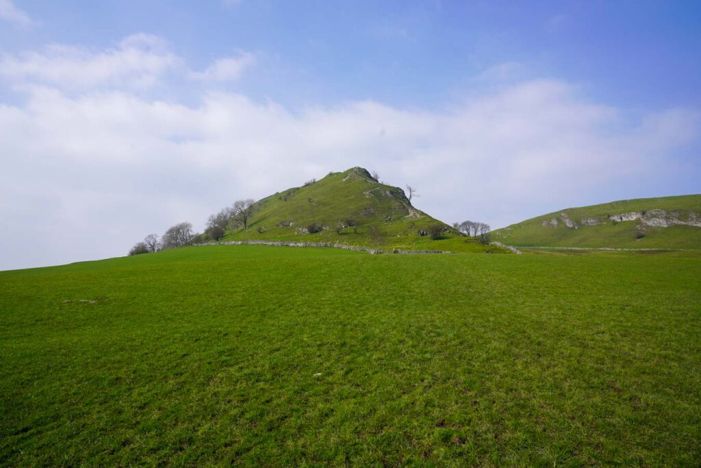 Grassy hillside leading up to the distinct peak of Chrome Hill in the Peak District Derbyshire with a worn walking path zigzagging to the summit under a partly cloudy sky. The open landscape and clear route highlight the experience of chrome hill walks across rolling green fields.