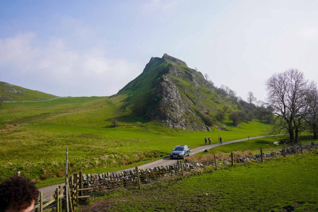 A car drives along a narrow country road beneath the steep ridge of Chrome Hill in the Peak District Derbyshire while a few hikers walk nearby. The sharp grassy and rocky slope rises dramatically above the landscape, highlighting a scenic route popular for chrome hill walks.
