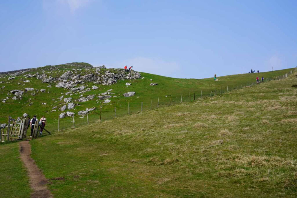 A grassy path leads through a gate toward a rocky hill in the Peak District Derbyshire as several hikers walk along the ridge ahead. The open landscape and gentle incline show an accessible section of chrome hill walks with wide countryside views.