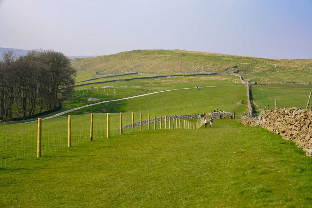 A wide grassy path bordered by fences and dry stone walls stretches across rolling hills in the Peak District Derbyshire with walkers in the distance. The gentle terrain and open views show an easier section of chrome hill walks through peaceful countryside.