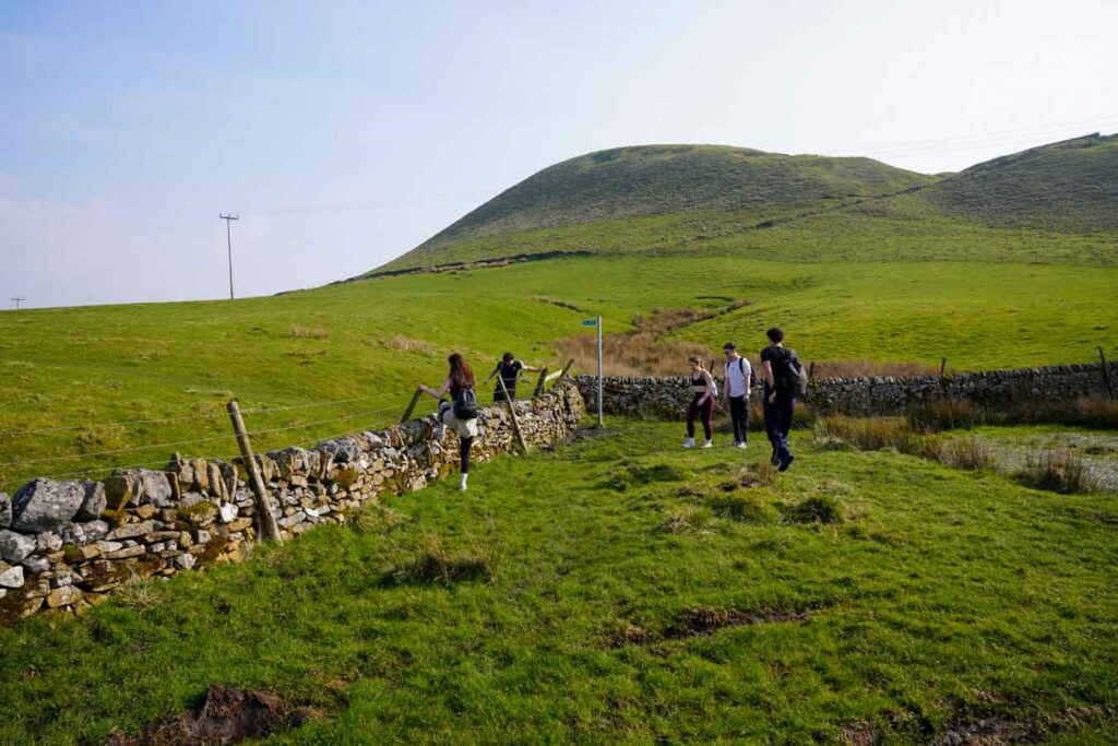 A group of hikers cross a stile over a dry stone wall in the Peak District Derbyshire with rolling green hills behind them. The rural path and open farmland setting show a typical access point along chrome hill walks.