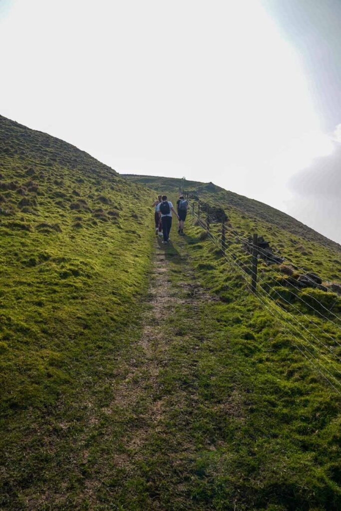 A group of hikers cross a stile over a dry stone wall in the Peak District Derbyshire with rolling green hills behind them. The rural path and open farmland setting show a typical access point along chrome hill walks.