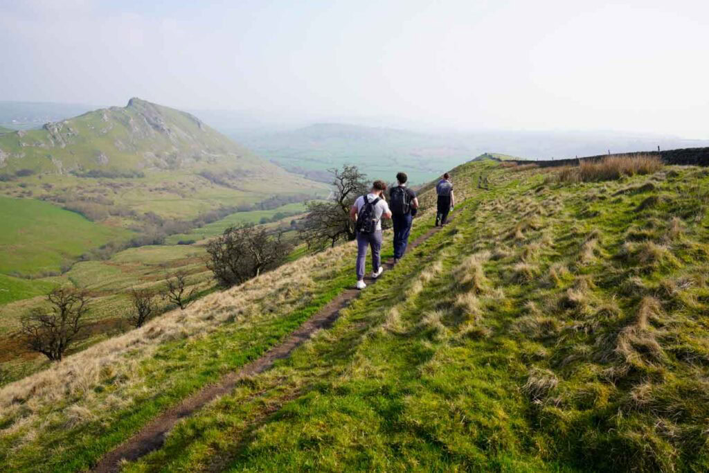 Three hikers walk along a narrow hillside path in the Peak District Derbyshire with sweeping valley views and Chrome Hill visible in the distance. The scenic ridge trail and elevated perspective capture the experience of chrome hill walks through rolling countryside.